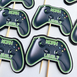 May include: Six green and black video game controller cupcake toppers with the name "Noah" and "Level 7" printed on them.