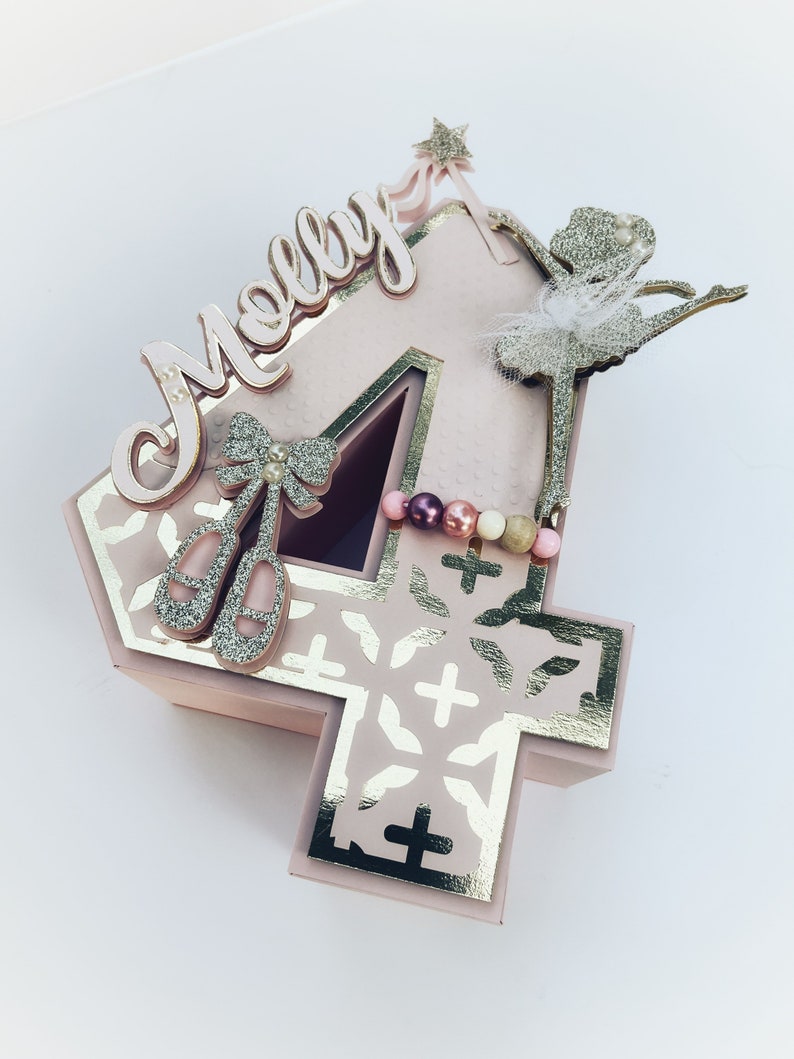 May include: A pink number four cake topper with a gold glitter ballerina, a pink bow with a pearl, and the name "Molly" in pink letters.