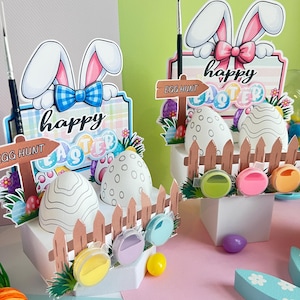 Può includere: Due decorazioni pasquali. Ognuna presenta una scatola bianca con una staccionata in legno, uova decorate e un cartello con la scritta "Buona Pasqua". Uno ha un papillon blu e l'altro un fiocco rosa. Sono inclusi dei pennelli.