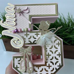 May include: Pink and gold number five cake topper with a ballerina, a bow, and ballet shoes. The topper is decorated with glitter and has the name "Mira" on it.