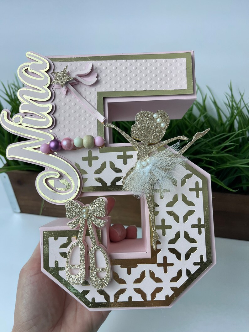 May include: Pink and gold number five cake topper with a ballerina, a bow, and ballet shoes. The topper is decorated with glitter and has the name "Mira" on it.