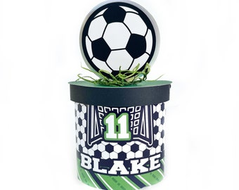 May include: A black and white soccer ball topper with green grass on a black and white soccer themed favor box with the number 11 and the name Blake.