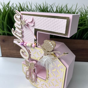 May include: A pink and gold number five cake topper with a ballerina cutout and the name "Viva" on the side. The number is decorated with a white tulle tutu, gold glitter, and pink and white beads.