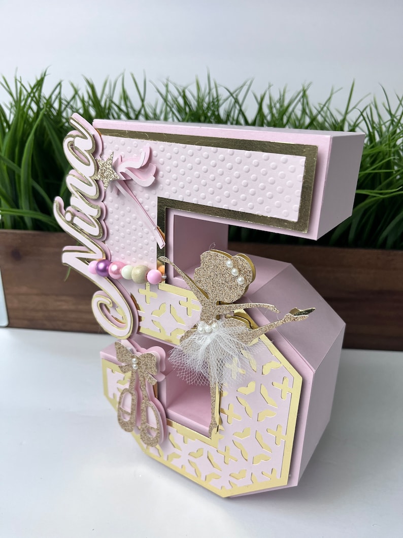 May include: A pink and gold number five cake topper with a ballerina cutout and the name "Viva" on the side. The number is decorated with a white tulle tutu, gold glitter, and pink and white beads.