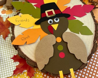 Puede incluir: Decoración de pavo de Acción de Gracias hecha a mano con papel marrón, amarillo, naranja, rojo y verde. El pavo lleva un sombrero negro con una hebilla dorada. El pavo tiene ojos saltones y las palabras "Dad", "friends" y "food" están escritas en las plumas.