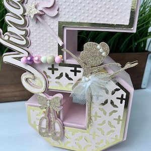 May include: Pink and gold number five cake topper with a ballerina figurine, a wand, and a bow. The number is decorated with a polka dot pattern and a gold trim.