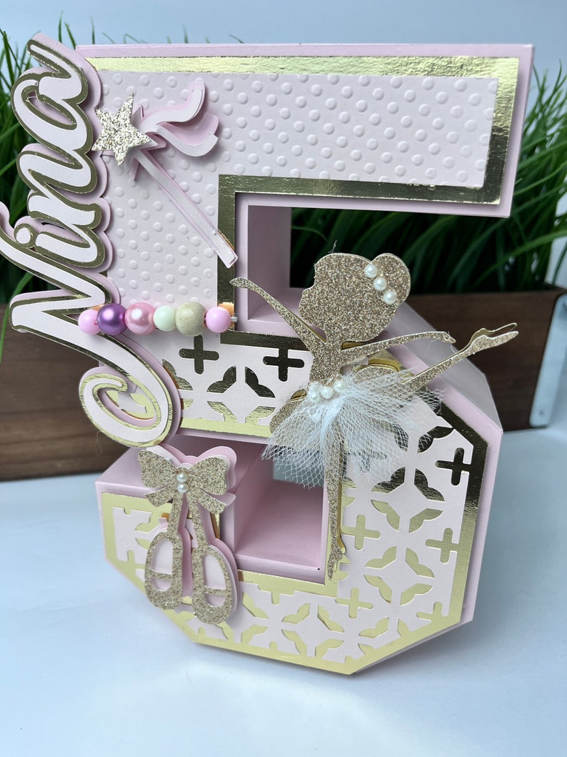 May include: Pink and gold number five cake topper with a ballerina figurine, a wand, and a bow. The number is decorated with a polka dot pattern and a gold trim.