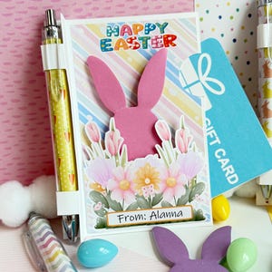 Könnte beinhalten: Eine Ostergrußkarte mit einer rosa Hasen-Silhouette und dem Schriftzug "Happy Easter". Die Karte ist mit Blumen und dem Text "From: Alanna" verziert. Eine blaue Geschenkkarte und bunte Stifte sind ebenfalls zu sehen.