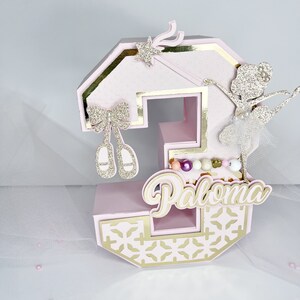 May include: Pink and gold number three cake topper with a ballerina, a wand, bows, and the name Paloma.