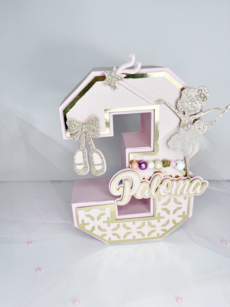 May include: Pink and gold number three cake topper with a ballerina, a wand, bows, and the name Paloma.