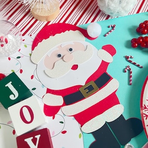 May include: A festive Christmas-themed arrangement featuring a Santa Claus cutout with glitter accents, candy canes, and wooden blocks spelling "JOY." The scene includes a small white Christmas tree and a fluffy white ornament, all set against a red and white striped background.