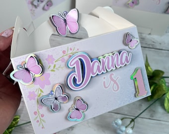 May include: White party favor boxes decorated with pink butterflies and the name "Danna" in a cursive font. The number "1" is also displayed. The boxes have a handle and are decorated with floral accents.