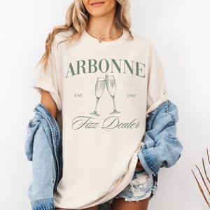 May include: A beige t-shirt with the text "Arbonne Est. 1980 Fizz Dealer" and a graphic of two champagne glasses.