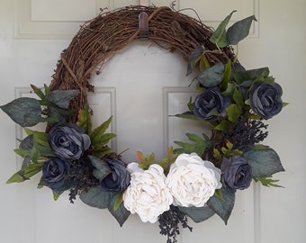Op de afbeelding: Een druivenrankenkrans versierd met witte en donkerblauwe kunstbloemen. De krans hangt aan een witte deur.