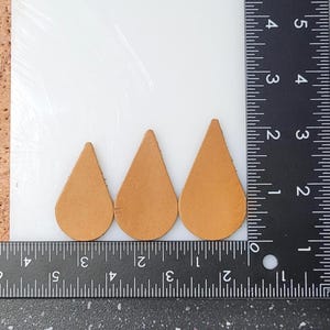May include: Three teardrop-shaped leather pieces in varying sizes, arranged from smallest to largest. The leather is a light tan color. A ruler with inch markings is placed next to the leather pieces for size comparison.