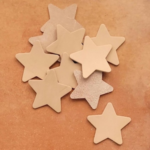 May include: A collection of light tan, five-pointed star-shaped cutouts. The stars are scattered on a brown surface. Some stars have a smooth finish, while others have a textured, suede-like appearance. These could be used for crafts or decorations.