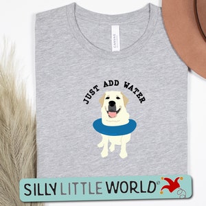 K&ouml;nnte beinhalten: Ein graues T-Shirt mit dem Text "Just Add Water" und einem Cartoon-Bild eines gelben Labradors, der eine blaue Schwimmweste tr&auml;gt. Der Text "Silly Little World" befindet sich am unteren Rand des Shirts.