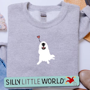 May include: A grey sweatshirt featuring a white dog illustration with a red heart above it. The text "SILLY LITTLE WORLD" is on a green and yellow banner below the dog.