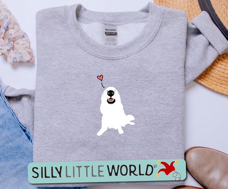 May include: A grey sweatshirt featuring a white dog illustration with a red heart above it. The text "SILLY LITTLE WORLD" is on a green and yellow banner below the dog.