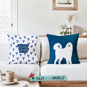 May include: Two decorative throw pillows on a white couch. One pillow has a blue and white heart pattern with the text "Just a girl who loves her dog". The other pillow is a solid dark blue with a white silhouette of a Great Pyrenees dog.