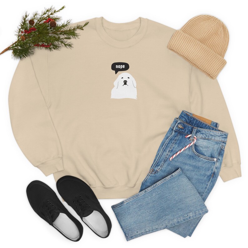 May include: A beige sweatshirt with a white dog illustration and the word "nope" in a speech bubble. The sweatshirt is laid out on a white background with a pair of black sneakers, blue jeans, and a beige beanie.