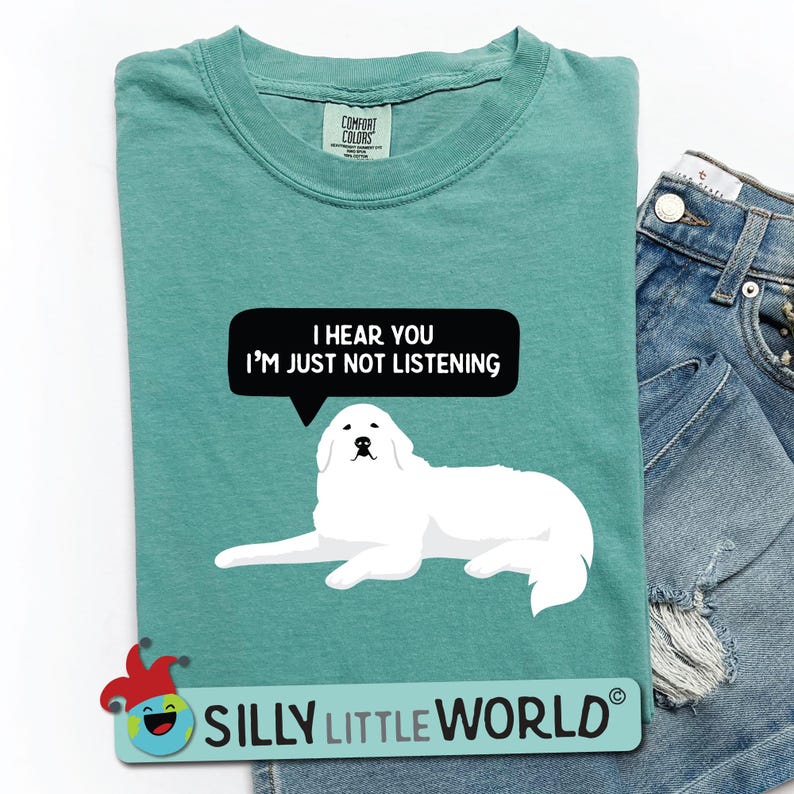 K&ouml;nnte beinhalten: Ein blaugr&uuml;nes T-Shirt mit einer wei&szlig;en Hundefigur und dem Text "I hear you I'm just not listening" in einer schwarzen Sprechblase. Der Text "Silly Little World" ist in Wei&szlig; auf einem roten und gelben Narrenhut-Motiv am unteren Rand des Shirts gedruckt.
