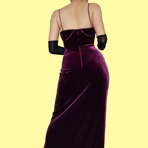 May include: A deep purple velvet gown with thin spaghetti straps and a fitted bodice. The dress has a long, flowing skirt and a zipper closure in the back. The model is wearing black gloves. The background is a solid yellow.