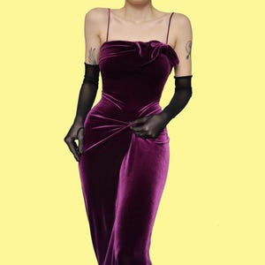 May include: A deep purple velvet gown with spaghetti straps and a bow detail at the bust. The dress features a fitted bodice and a flowing skirt. The model is wearing sheer black gloves. The background is a solid yellow.