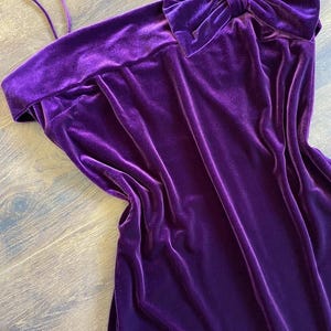 May include: A vibrant purple velvet dress with a bow detail. The dress features a strapless design with a wide neckline and a large bow accent on the shoulder. The fabric has a soft, luxurious texture.