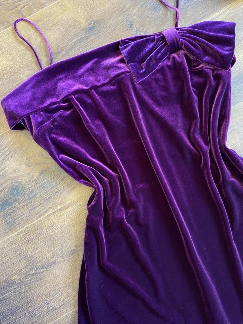 May include: A vibrant purple velvet dress with a bow detail. The dress features a strapless design with a wide neckline and a large bow accent on the shoulder. The fabric has a soft, luxurious texture.