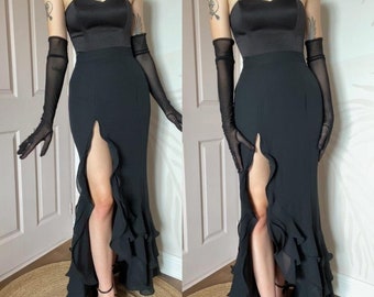 May include: A black strapless evening gown with a sweetheart neckline and a high slit. The bodice is satin, and the skirt features cascading ruffles. Long, sheer black gloves complete the look. The dress is paired with black strappy heels.