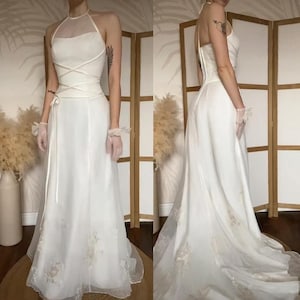 Puede incluir: Un vestido de novia blanco con cuello halter y falda fluida. El corpiño presenta detalles entrecruzados y un panel transparente. La falda está adornada con bordados florales cerca del dobladillo. El vestido es largo y tiene una cola.