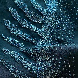 May include: Close-up of a dark blue fabric with a pattern of silver and blue beads. The beads are arranged in a geometric design, creating a shimmering effect.