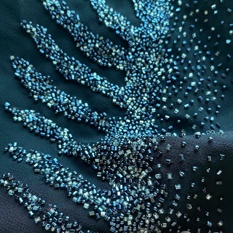 May include: Close-up of a dark blue fabric with a pattern of silver and blue beads. The beads are arranged in a geometric design, creating a shimmering effect.