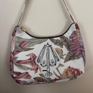 May include: A white shoulder bag with a floral pattern in shades of burgundy, green, and yellow. The bag features a black zipper and a black outline. A black and white lamp graphic is centered on the front.