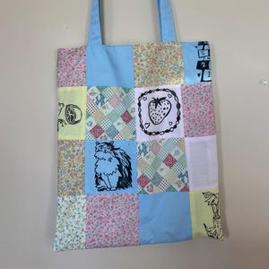 Op de afbeelding: Een patchwork tote bag met een lichtblauwe handgreep en body. De tas heeft een verscheidenheid aan stoffen vierkanten in pastelkleuren en patronen, waaronder bloemenprints, een aardbei, een kattenillustratie en een citroen.