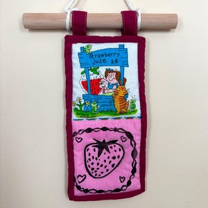 Op de afbeelding: Een wanddecoratie met een aardbeienthema, met een sapkraamscène en een aardbeienillustratie. Het kunstwerk heeft een bordeauxrode rand, een houten deuvel en een wit koord om op te hangen. Afmetingen in cm.