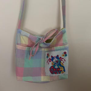 Op de afbeelding: Een pastelkleurige schoudertas met een kattenapplicatie. De tas heeft een lichtroze, blauw, geel en groen ruitpatroon. Op de voorkant is een patch met een kattenontwerp genaaid. De tas heeft een lange riem en een trekkoordsluiting.
