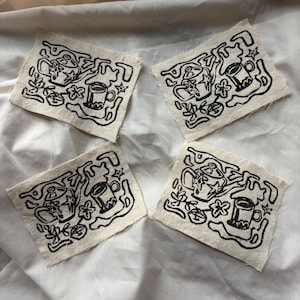 May include: Four square fabric patches with a hand-drawn black design on a cream background. Each patch features a coffee cup, floral elements, and abstract shapes. The patches are arranged on a white fabric surface.