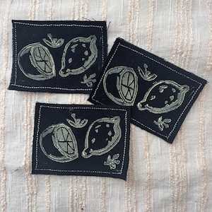 May include: Three black fabric patches with a lemon design. Each patch features a lemon and a lemon slice in a light green color, with white stitching around the edges. The patches are arranged on a textured, off-white fabric.