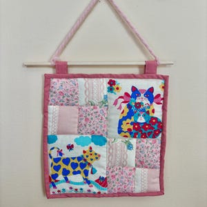 May include: A handmade wall hanging featuring a patchwork design with pink floral and solid fabric squares. The center showcases two cat illustrations and a cow illustration with heart-shaped spots. A pink rope and wooden dowel are used for hanging.