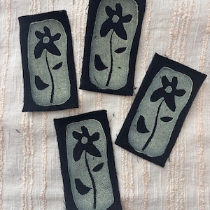 May include: Four black fabric patches with a floral design. Each patch features a light green flower with a stem and leaves, set against a black background. The patches are rectangular and have a raw edge.