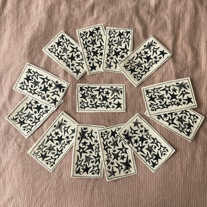Könnte beinhalten: Ein Satz von zehn Karten mit einem schwarzen Sternenmuster auf cremefarbenem Hintergrund. Die Karten sind in einem Kreis auf einer hellbraunen Stoffoberfläche angeordnet.