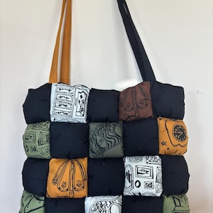 Op de afbeelding: Handgemaakte tote bag met een patchwork ontwerp. De tas heeft een raster van zwarte vierkanten afgewisseld met vierkanten in verschillende kleuren en patronen, waaronder klokken, paraplu's en architectonische tekeningen. De bandjes zijn bruin en zwart.