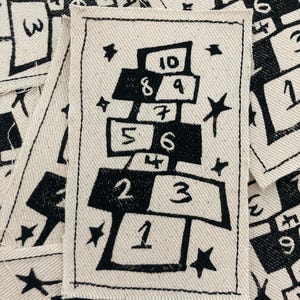 Op de afbeelding: Een zwart-witte illustratie van een tikkertje met de nummers 1 tot en met 10. De illustratie is gedrukt op een witte stoffen patch.