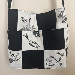 Op de afbeelding: Een zwart-wit geblokte crossbodytas met een lange riem. De tas heeft een herhalend patroon van visillustraties op de witte vierkanten. De tas heeft een voorvak en een zwarte riem.