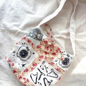 Op de afbeelding: Een patchwork tote bag met een witte band. De tas heeft vierkanten met zwart-witte illustraties van een appelklok, sterren en kledingstukken. De achtergrond heeft een bloemenpatroon met rode bloemen.