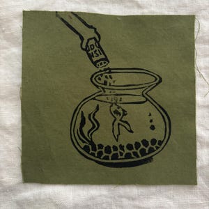 Op de afbeelding: Olijfgroene stoffen patch met een zwart-wit illustratie. Het toont een hand die vloeistof uit een fles in een vissenkom giet met een vis en stenen.