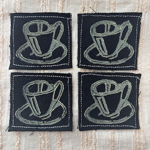 Könnte beinhalten: Vier quadratische schwarze Stoffaufnäher mit einem hellgrünen Kaffeetassen-Design. Jeder Aufnäher hat einen weiß genähten Rand. Die Aufnäher sind auf einem cremefarbenen, strukturierten Stoff angeordnet.