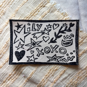 May include: A rectangular patch with a black border features a hand-drawn design. The design includes stars, hearts, the word "Lily", and the letters "XOXO" in black on a cream-colored background. The patch is on a textured, cream-colored fabric.